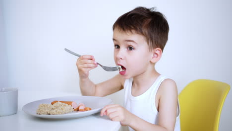 young child enjoys meal at home. a boy in a white tank top sits at a table with a plate of rice and sausages, thoughtfully eating his meal