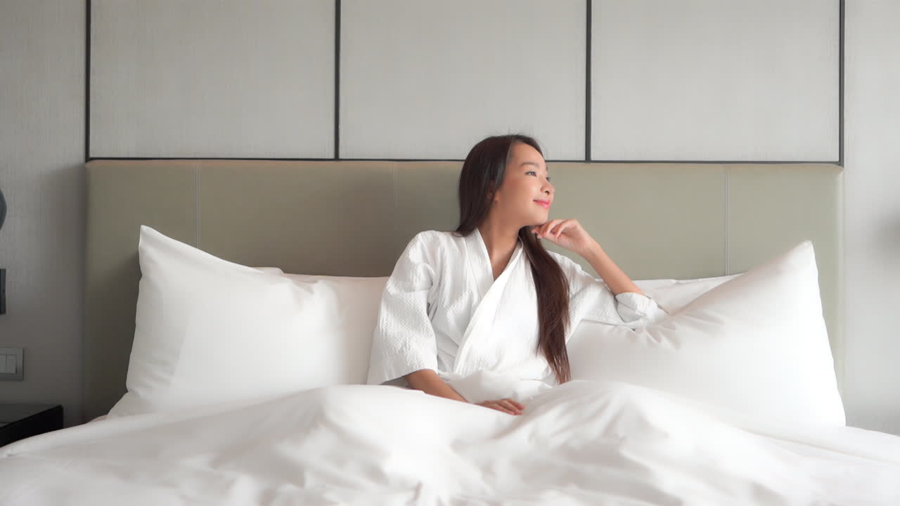 Sitting up in a huge hotel suite bed smiles as she looks out toward the light of the window