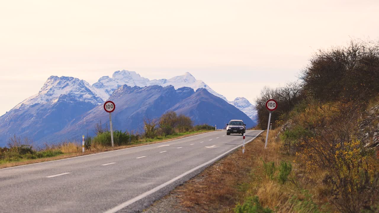 A car travels along a picturesque road with snow-capped mountains in Glenorchy, New Zealand. Clear skies and natural beauty enhance the scene