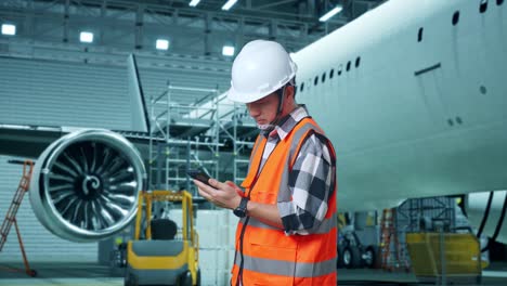 vista lateral de un ingeniero masculino asiático con casco de seguridad usando un teléfono inteligente mientras está de pie con el avión en el hangar