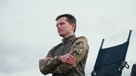 from the side a confident man with a short haircut in an army camouflage uniform stands confidently against the sky and looks to the side with his arms folded on his chest outside the city