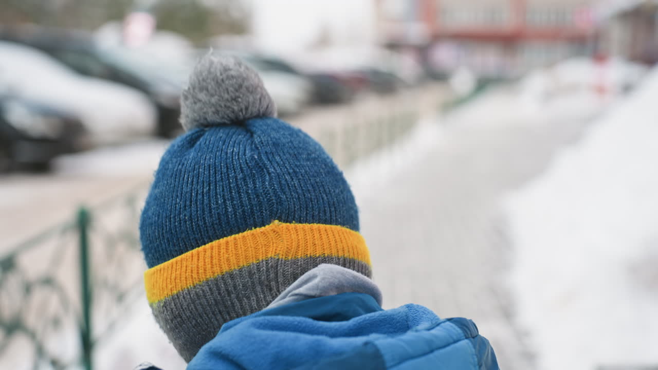 雪の歩道を母親の隣で歩く息子。黄色いストライプとグレーのポンポンが付いた青いビーニー帽の後ろ姿のクローズアップ。ウールの質感が快適な冬のディテールを際立たせ、遊び心のある子供時代の瞬間、都会の散歩。