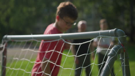 a ball is shot into a goal post as the goalkeeper in red misses the save and adjusts his glasses, with blur view of two person standing in the background