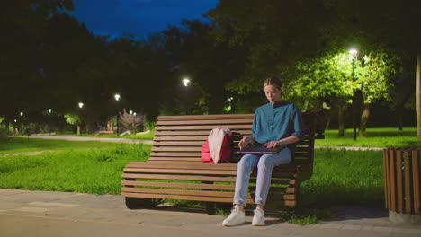 una joven se sienta en un banco del parque al amanecer, abriendo una computadora portátil con una bolsa roja a su lado en un entorno al aire libre tranquilo, el fondo presenta árboles, postes de luz, vegetación, personas paseando y un cielo azul profundo