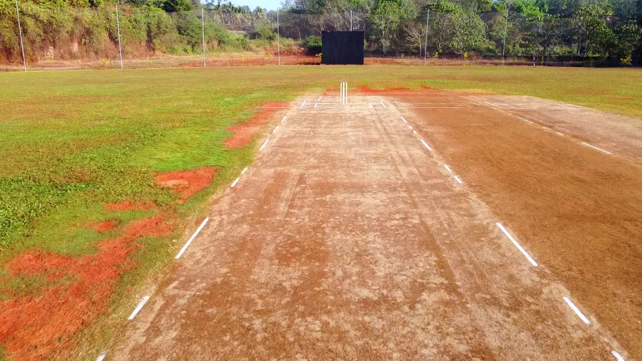 Ripresa aerea con drone di un campo da cricket rurale e dei wicket in una giornata di sole, che mostra la linea di battuta in terra battuta, il campo esterno verde e gli alberi tropicali circostanti in un'ambientazione tranquilla.