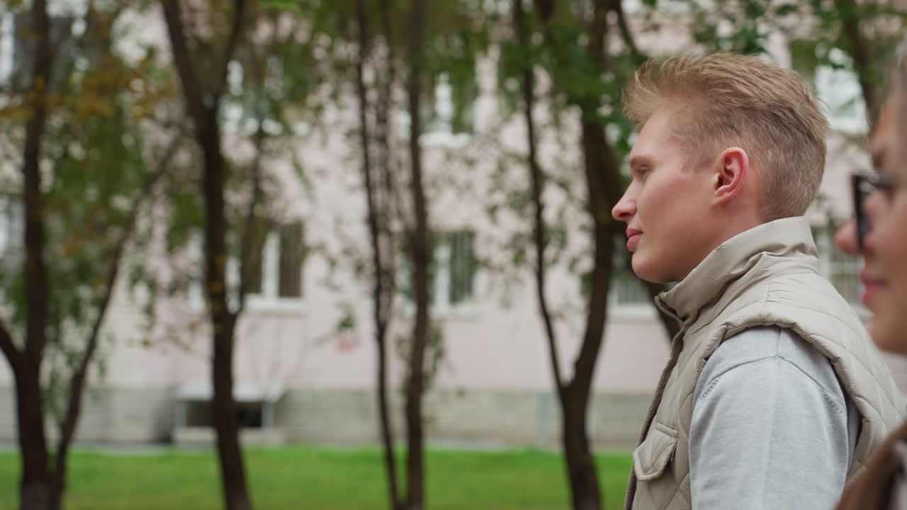 Lovely friends walking side by side outdoors as woman glances at man with soft smile and hair swaying in wind, man stays focused ahead, trees and buildings visible in background