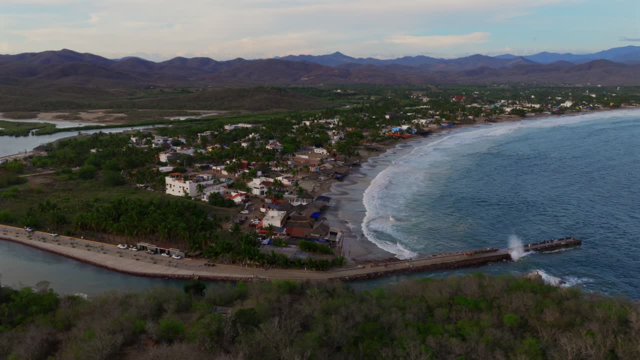 High aerial view of pier extending into the ocean, small village along beach, and lush coastal terrain in Punta Perula, Costalegre, Mexico