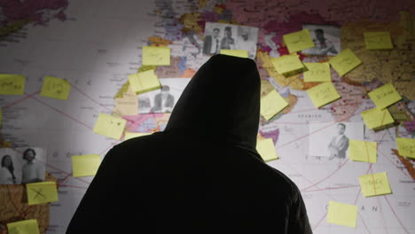 back view of unrecognizable man in hood making notes on paper stickers stuck to investigation map and searching connections