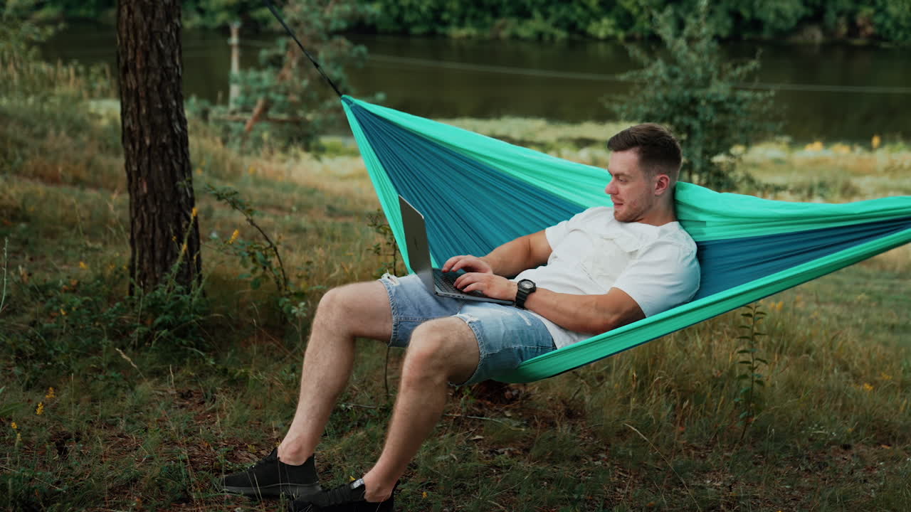 Muscular Caucasian man half-lies in a hammock and works on his laptop. Focused man typing a message. Nature backdrop.