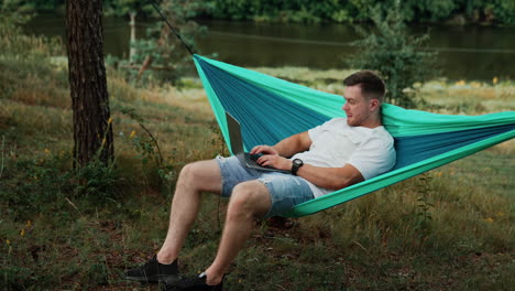 muscular caucasian man half-lies in a hammock and works on his laptop. focused man typing a message. nature backdrop.