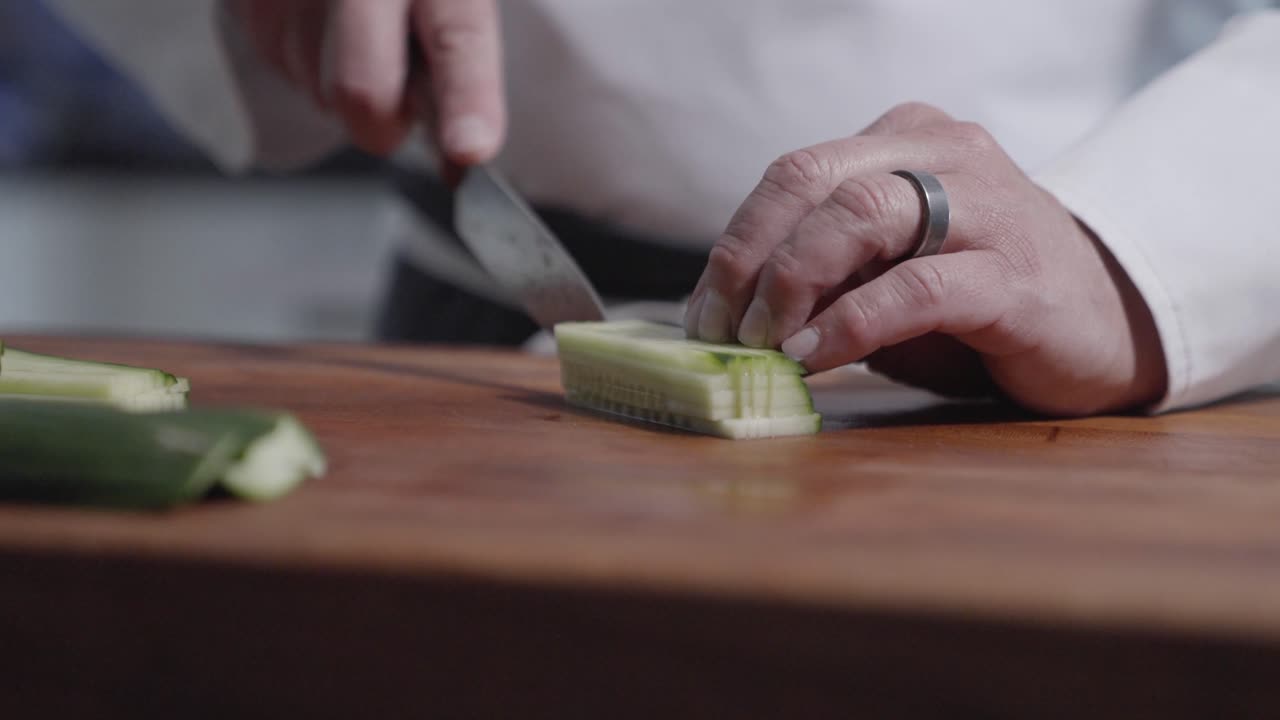 “Close-up static shot of a chef chopping and dicing cucumbers into small squares on a wooden cutting board before walking away. Highlights precise knife skills and culinary preparation.”