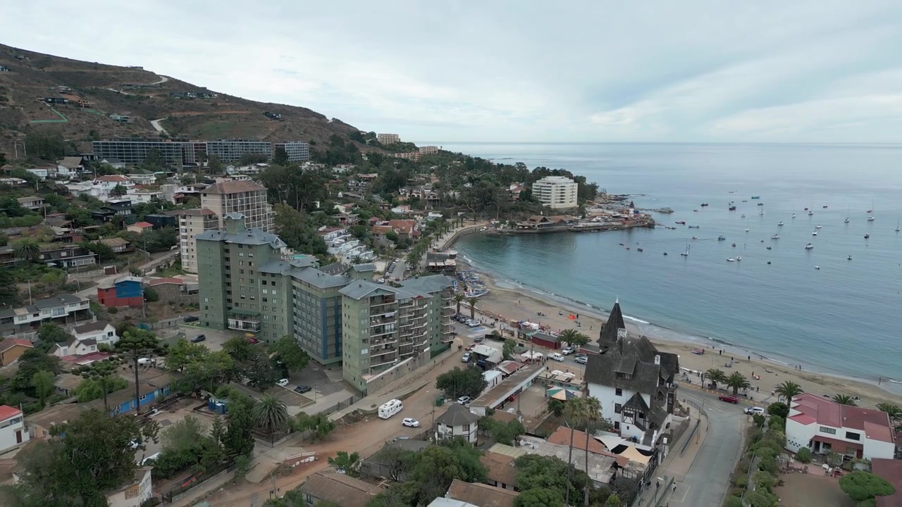 Volo in allontanamento sopra la città di Papudo che rivela condomini, spiaggia sabbiosa, baia calma con barche da pesca ancorate e colline aride che si ergono dietro sotto un cielo nuvoloso. Papudo, Valparaíso, Cile.