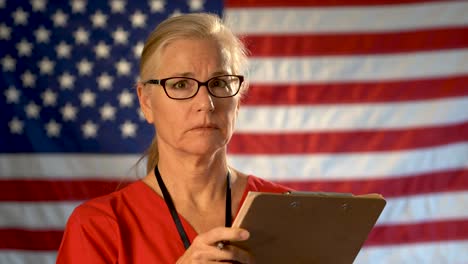 Medium-tight-portrait-of-concerned-healthcare-nurse-looking-at-clipboard-and-then-at-camera-with-concerned-look-on-her-face-with-American-flag-behind-her