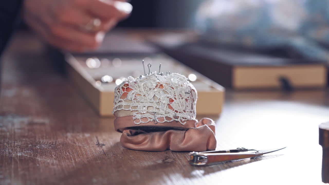 Sewing pins in a white lace sponge for dressmaker on the wooden table. Nice pincushion on the table on the background of tailor hands working in workshop. Close-up.