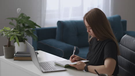 Junge-Studentin-Lernt-Zu-Hause,-Sieht-Sich-Schulungsvideos-Auf-Dem-Laptop-Bildschirm-An-Und-Macht-Sich-Notizen-Im-Notizbuch,-Fernunterricht-Während-Der-Coronavirus-Pandemie