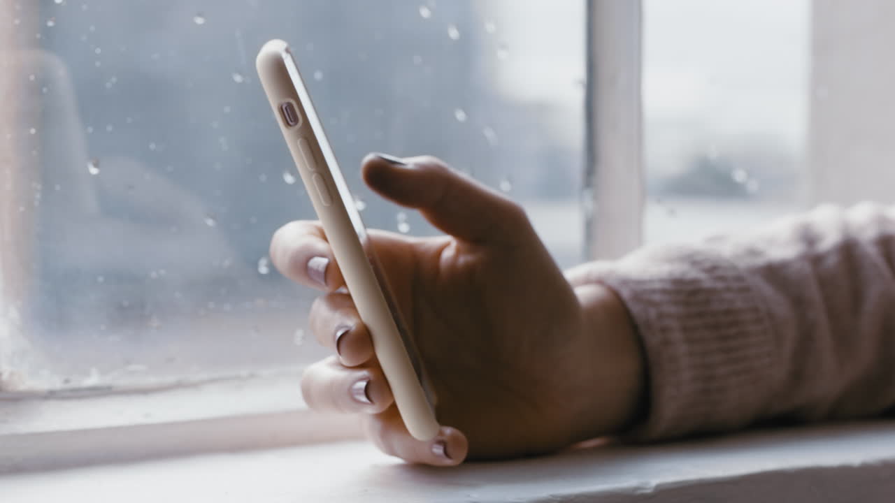close up woman hand using smartphone browsing online messages reading social media enjoying mobile communication standing by window relaxing at home on cold rainy day