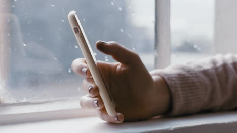 Cerrar-La-Mano-De-Una-Mujer-Usando-Un-Teléfono-Inteligente-Navegando-Mensajes-En-Línea-Leyendo-Las-Redes-Sociales-Disfrutando-De-La-Comunicación-Móvil-De-Pie-Junto-A-La-Ventana-Relajándose-En-Casa-En-Un-Día-Frío-Y-Lluvioso