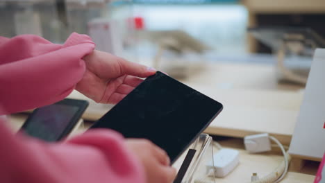 lady picks up tablet from its stand in electronics store, tablet is connected with electric cord, while other tablets are neatly arranged on the table, holding it gently as she observes the screen