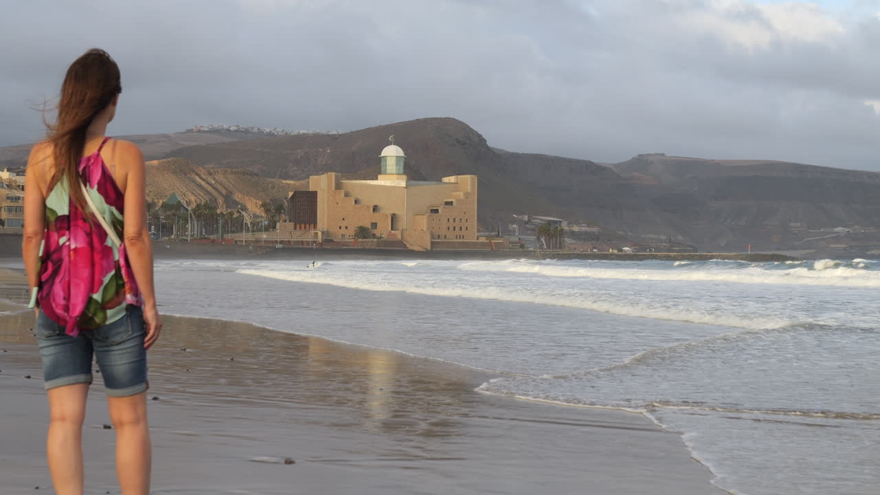 Ripresa cinematografica di una donna che ammira l'auditorium Alfredo Kraus mentre le onde si infrangono sulla riva della spiaggia di Las Canteras, sull'isola di Gran Canaria.