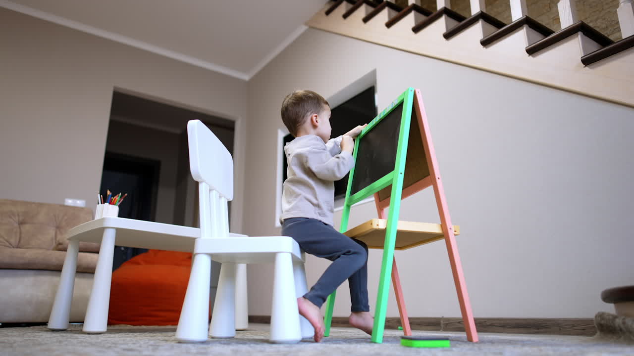 Young child drawing at a colorful easel. A child engages in drawing on a green and pink easel while sitting on a chair in a cozy living room