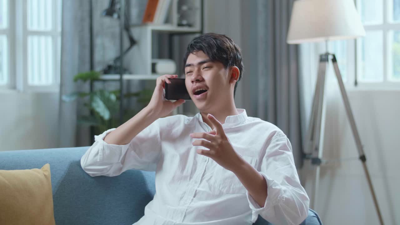 Smiling Asian Man Answering The Call On Smartphone While Lying On Sofa In The Living Room