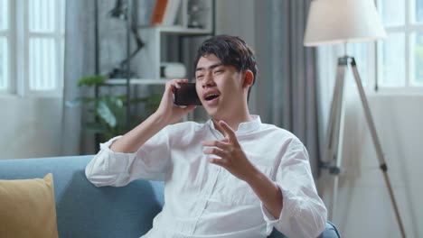 smiling asian man answering the call on smartphone while lying on sofa in the living room
