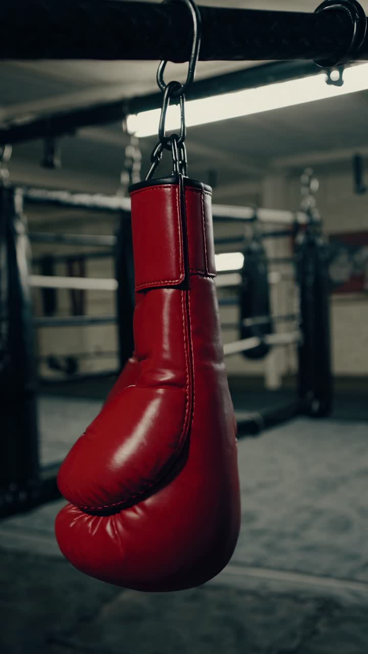 Close-up video of red boxing gloves hanging in a dimly lit gym, shot from a low angle