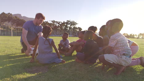 niños pequeños de la escuela y el maestro sentados con la pelota en el campo