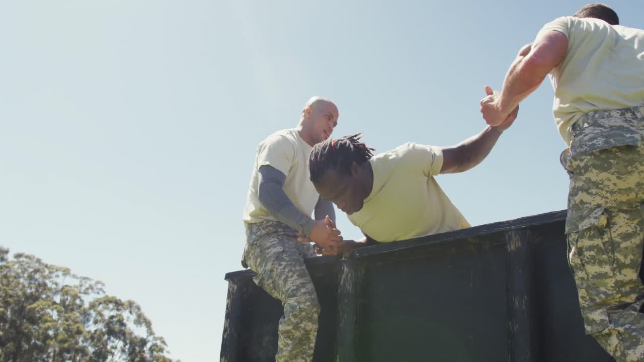 Three diverse fit male soldiers helping another climb high hurdle on army obstacle course