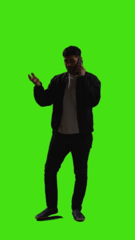 Vertical-Video-Full-Length-Shot-Of-Young-Man-Talking-On-Mobile-Phone-Standing-Against-Green-Screen-With-Low-Key-Lighting-1