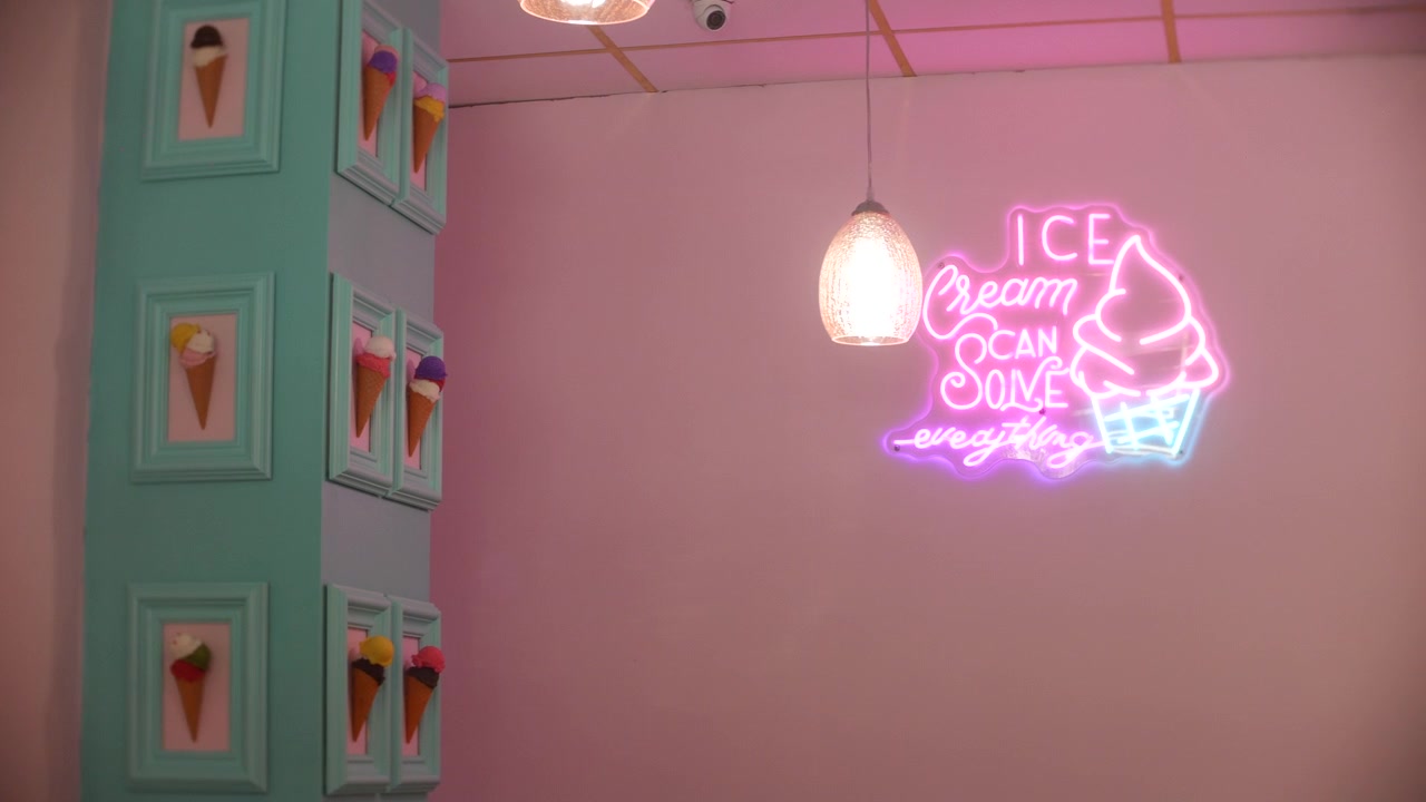 Letreiro de sorvete neon com luzes rosa e azuis brilhando em uma parede pastel de sorveteria, ao lado de decorações de cones emolduradas. Lâmpadas penduradas e acabamento turquesa criam um interior colorido e retrô de uma loja de sobremesas.