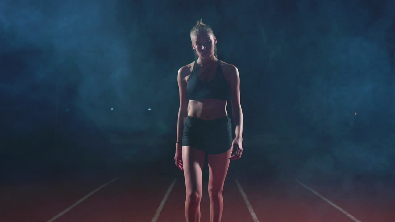 una delgada joven atleta pasa a través del humo, mira a la cámara y se acerca a la línea de salida de la carrera