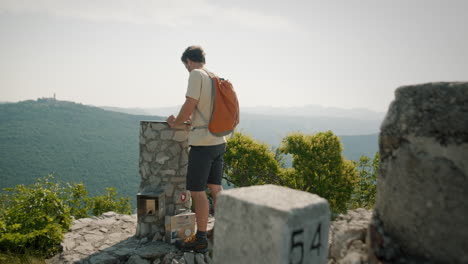 hiker with an orange backpack on the top of mountain saboti standing by a rock monumet with a compass plate for direction of cities or places