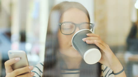 camera focuses on a brunette woman in glasses through the window drinking coffee and texting on the smartphone while she is sitting at a table in a cafe