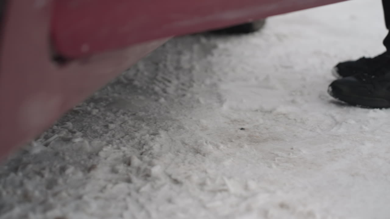 Close up side view of person wearing black boots walking across snow covered ground toward car door opening with flakes falling and parked cars visible in background during cold winter day