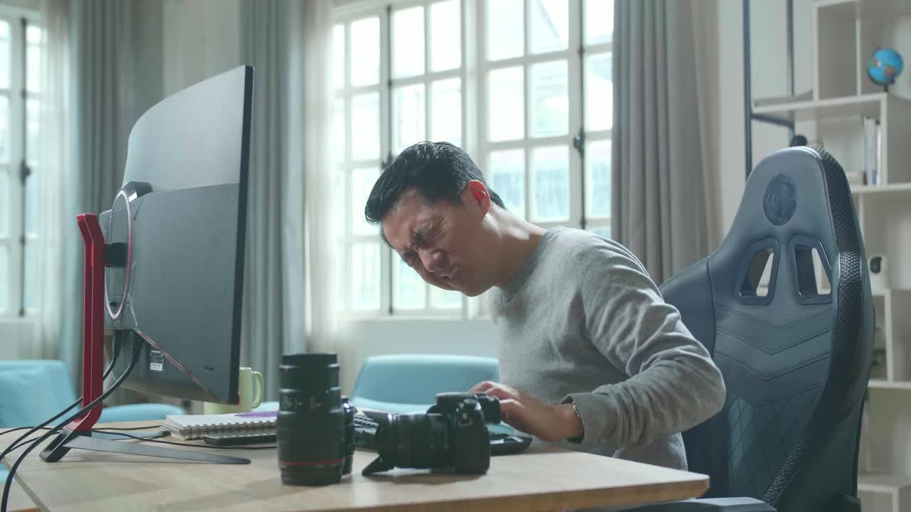 Asian Cameraman In Long Sleeved T-Shirt Having Backache While Using Desktop Computer For Working At Home.