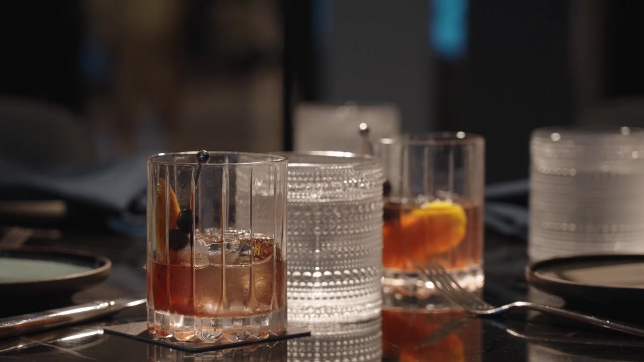 Filmato 4K di un cocktail Old Fashioned servito con guarnizione di mirtillo e scorza d'arancia in un elegante ristorante di lusso con atmosfera suggestiva, che cattura la raffinata mixology e l'atmosfera cinematografica.