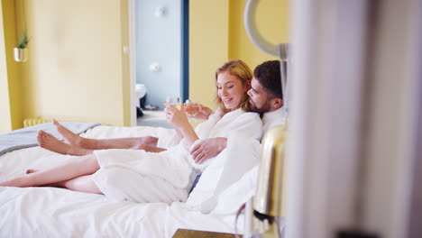 Mixed-race-young-adult-couple-reclining-on-the-bed-in-a-hotel-room-in-bathrobes-holding-glasses-of-wine-and-talking,-handheld-shot-moving-into-room-from-doorway