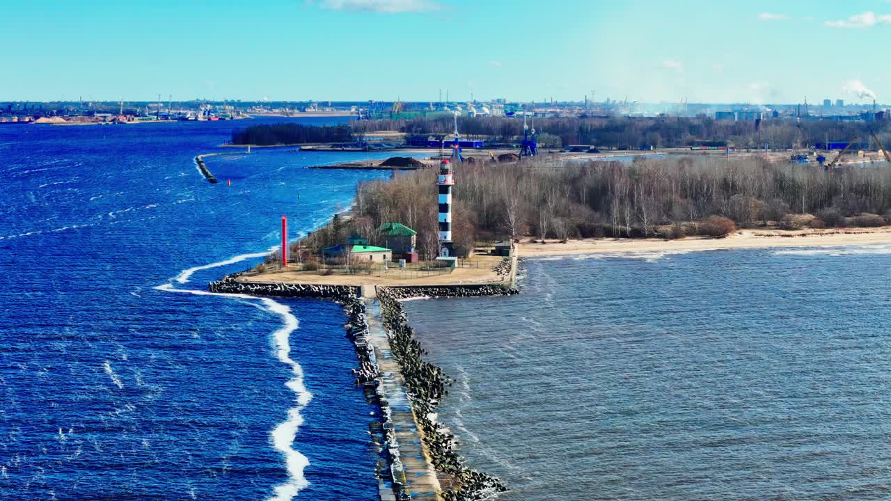 A striking black-and-white lighthouse rises from a long stone jetty, dividing vivid blue waves and a tranquil sandy shoreline backed by leafless trees.