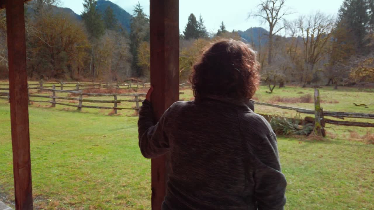 A lone middle aged woman relaxing on a cabin porch on a lush green rustic farm in a rain forest