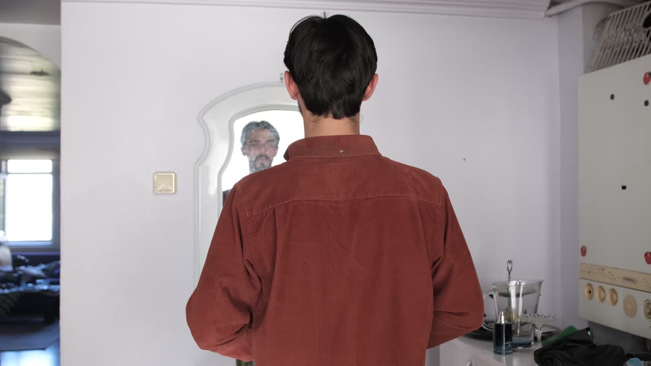 Young caucasian bearded man wearing his shirt in front of the mirror at home, people look in the mirror to fix themselves