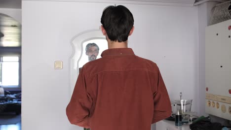 young caucasian bearded man wearing his shirt in front of the mirror at home, people look in the mirror to fix themselves