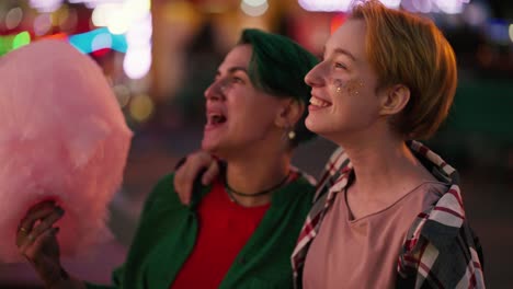 Happy-blonde-girl-with-glitter-on-her-face-in-a-checkered-pink-t-shirt-and-a-girl-with-short-green-hair-eating-a-large-pink-cotton-candy-and-looking-at-the-bright-rides-in-the-amusement-park-during-their-LGBT-date-in-the-evening