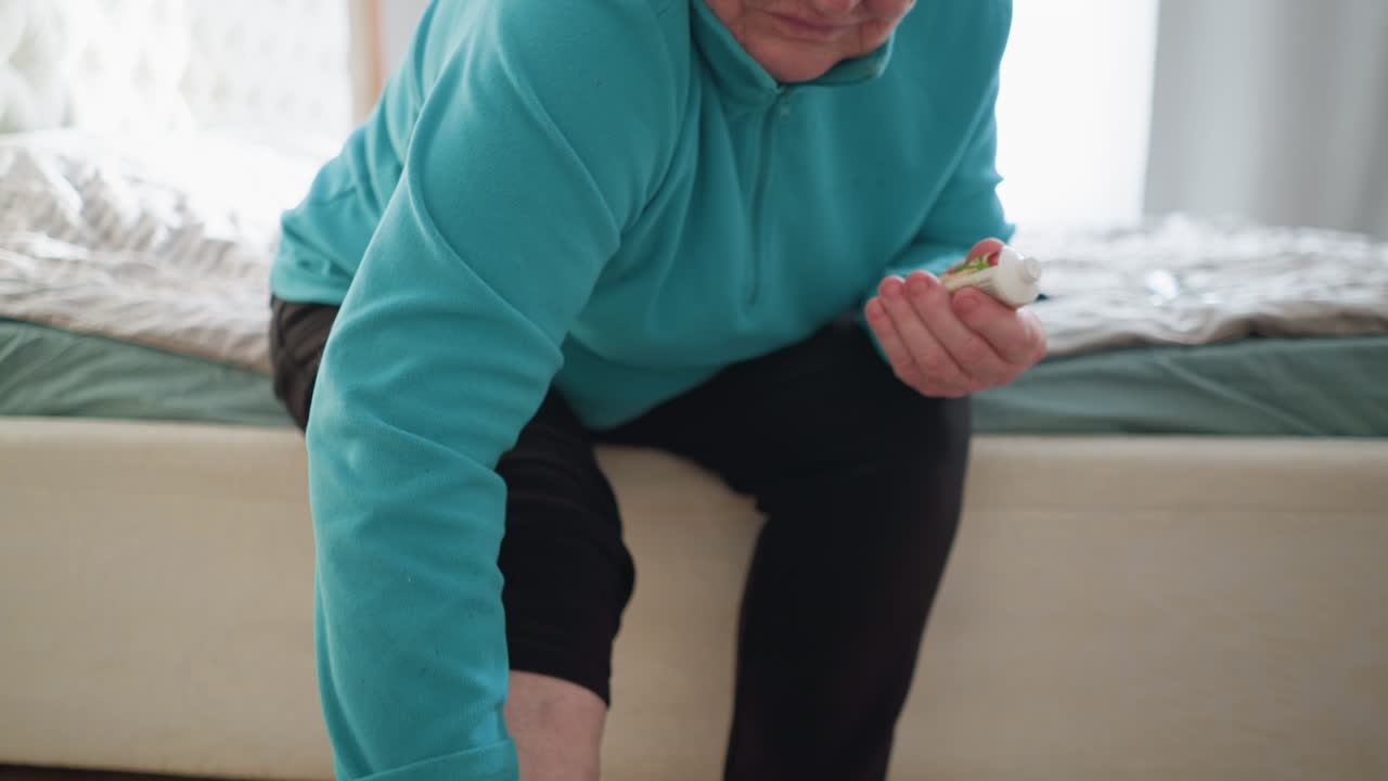 Old woman in green sitting on bed, reaching down to touch her foot. One sock on, one off. A moment of self-care, as she applies treatment to her foot in a cozy, peaceful bedroom setting with soft light