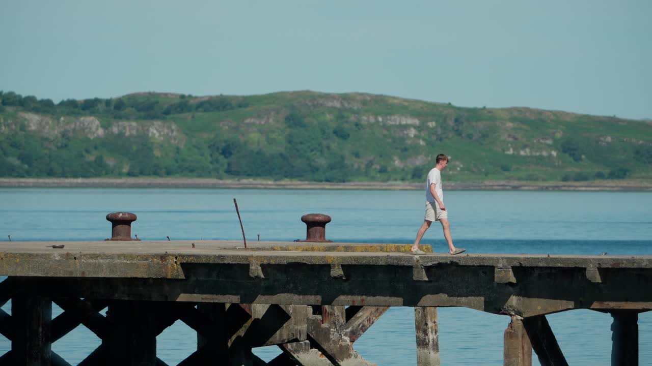 Young man strolls along an old pier over calm water, framed by distant shoreline and lush hills—evoking peaceful solitude and coastal reflection