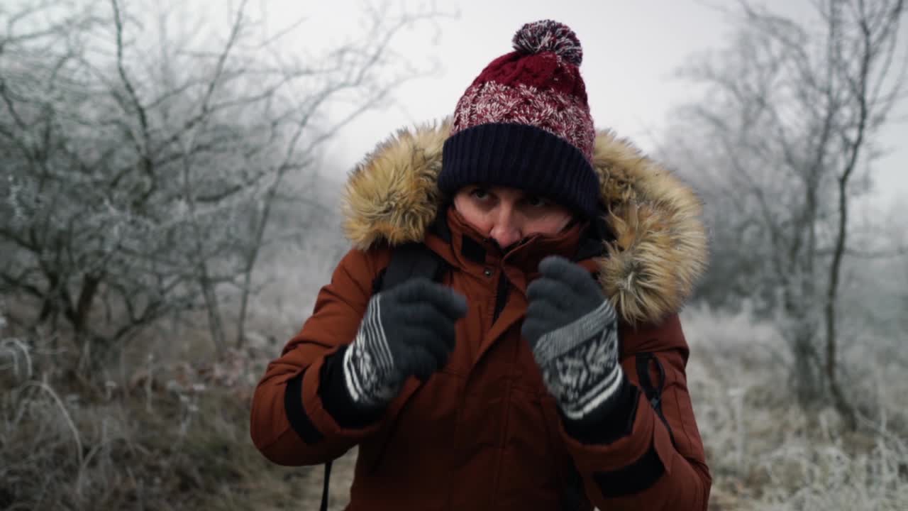 hombre con sombrero de algodón parado entre árboles blancos congelados y cubriendo su rostro con abrigo de invierno marrón