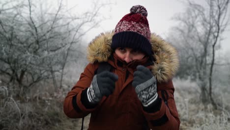 Man-with-cotton-hat-standing-between-frozen-white-trees-and-covering-his-face-with-brown-winter-coat