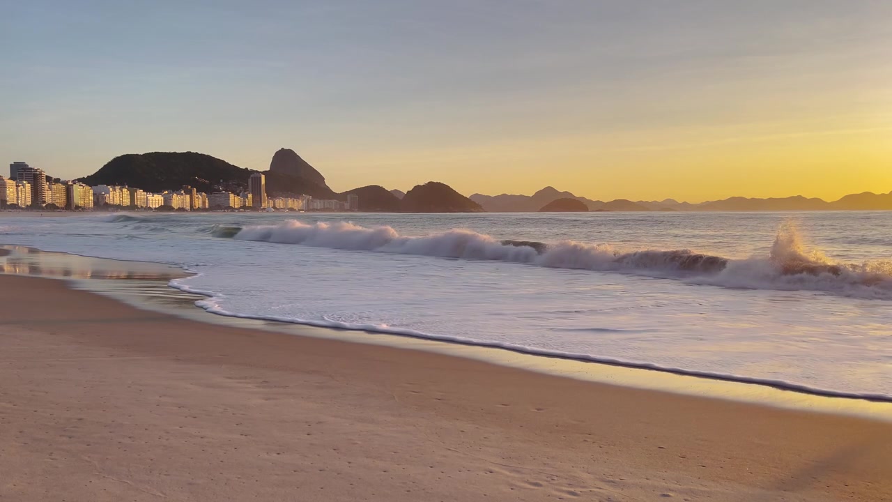 Plano geral estático na hora dourada do nascer do sol sobre a Praia de Copacabana com ondas suaves, silhueta do Pão de Açúcar e horizonte do Rio em luz quente e brilhante