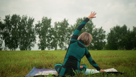 back view woman with grey hair lying on one side of yoga mat practicing side bend pose, arms extended, rising slowly in serene grassy field under cloudy skies with trees in the distance