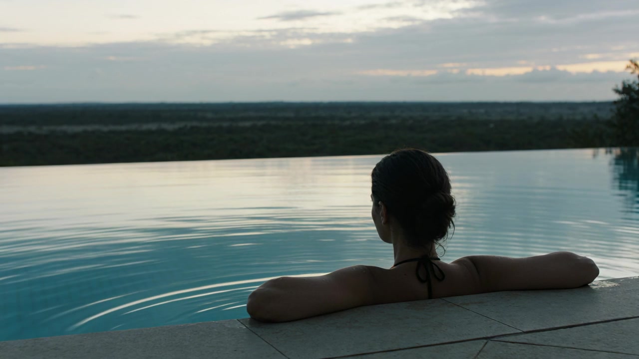 Ruhende Frau im Neckholder-Top bewegt die Arme, erzeugt Wellen am Infinity-Pool-Rand und beobachtet die Dämmerung.